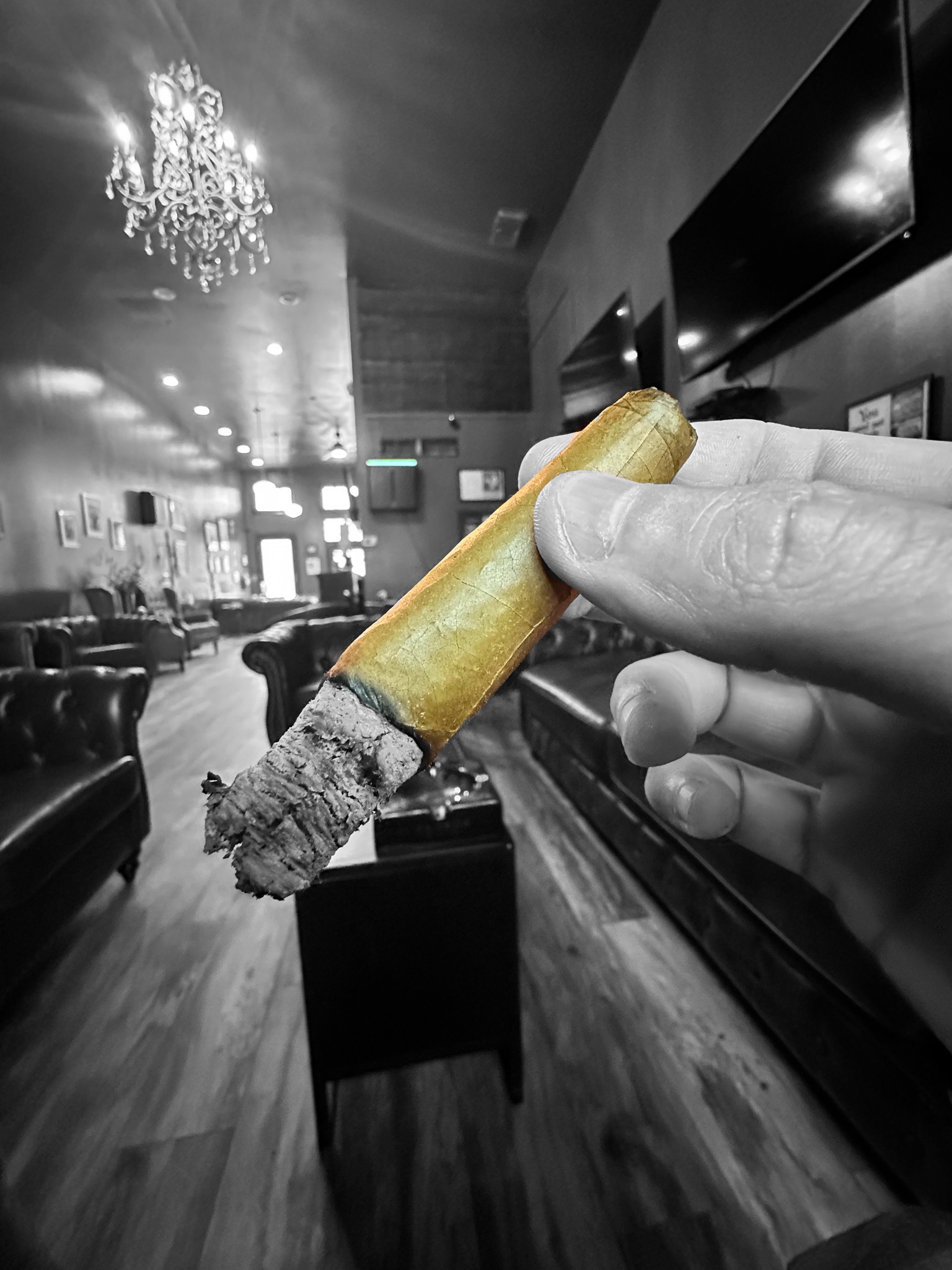 Close-up of a person's hand holding a lit cigar with an ash tip, in selective color highlighting the golden wrapper, against a black and white background of a lounge with leather sofas and a chandelier.