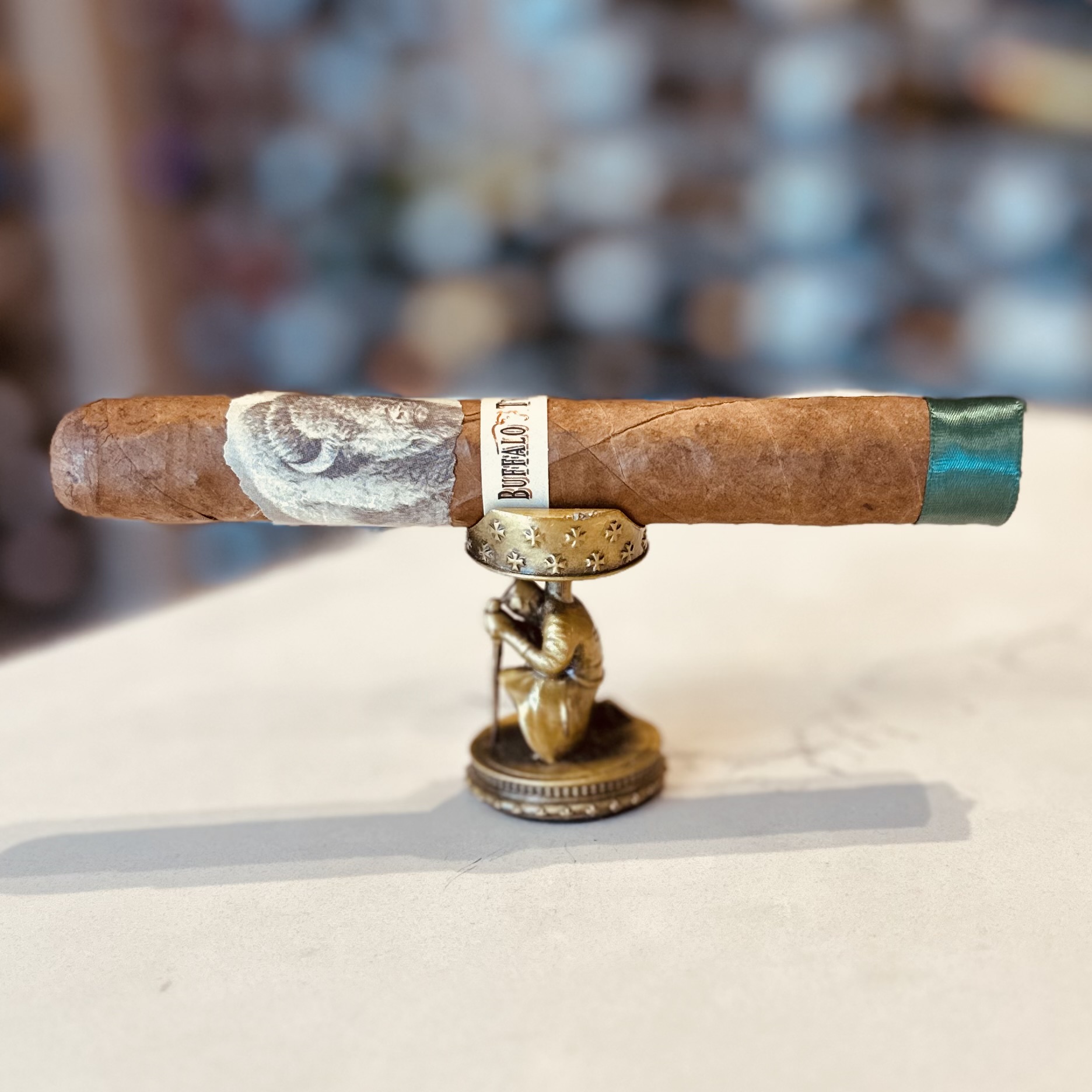 A premium cigar with a rich, tan wrapper and a distinctive band displaying the name 'Buffalo Trace'. The cigar is perched on an ornate, bronze-colored statue of a sitting figure, possibly a classical character, with a blurred background that hints at a cozy, well-stocked cigar lounge.