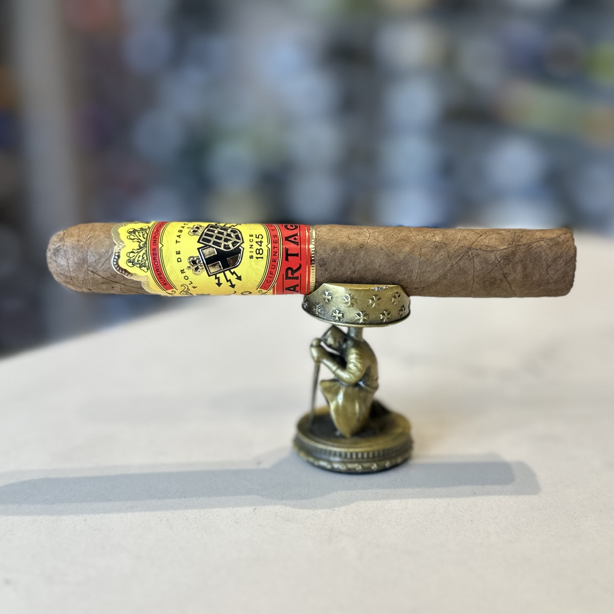 The image shows a cigar resting horizontally on a small stand. The cigar has a rich brown wrapper with a slightly oily sheen, indicating it may be a quality, hand-rolled product. Around the cigar are two bands; the larger one near the head of the cigar is primarily yellow with red and black accents, featuring a coat of arms and the text "PARTAGAS" in bold letters. Below this band is a narrower one with a patterned design. The stand appears to be a small, bronze-colored figurine of a person kneeling, adding an artistic touch to the presentation of the cigar. The background is blurred, drawing focus to the cigar and stand in the foreground.