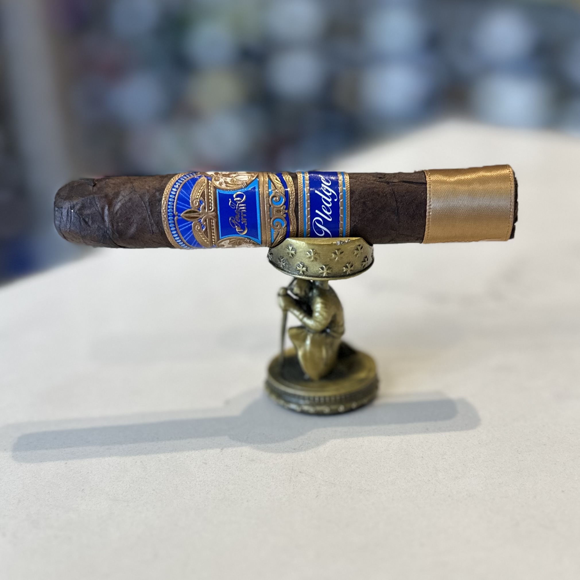 A close-up image of a richly colored E.P. Carrillo Pledge cigar with a prominent blue and gold label, resting on a metallic ring stand featuring a small figurine. The cigar is set against a soft-focus background, highlighting its detailed construction and the elegant, embossed branding on the label.