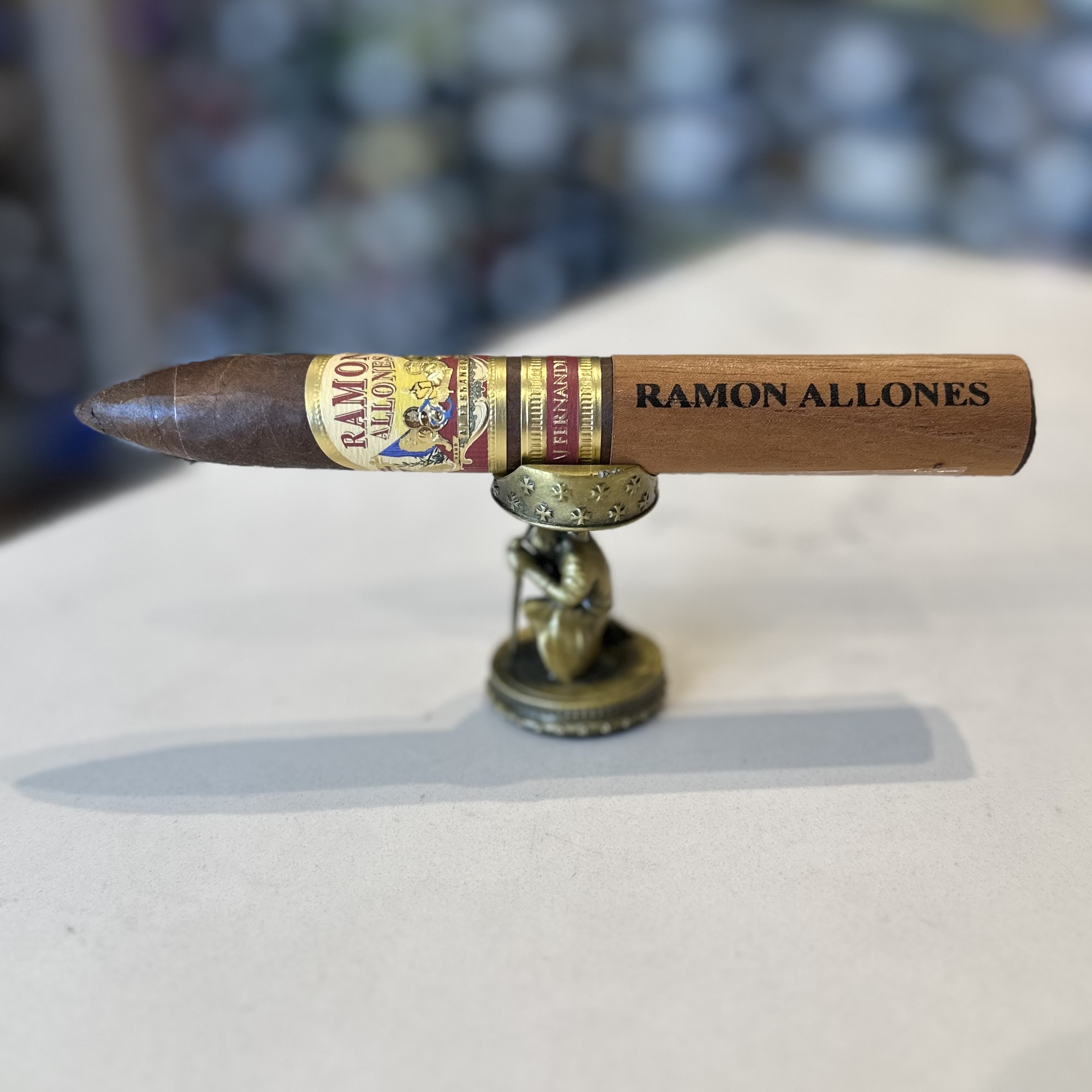 The image features a premium cigar perched on a small, decorative metal stand. The cigar has a glossy, ornate band with the inscription "RAMON ALLONES AJ FERNANDEZ" in a mix of elaborate and bold fonts, set against a golden background with red accents and decorative elements that suggest luxury and tradition. The cigar itself has a rich, chocolate brown wrapper, indicative of a full-bodied flavor profile. The background is softly blurred, drawing attention to the detailed cigar band and the craftsmanship of the cigar. The metal stand has a circular base and is adorned with intricate designs, contributing to the overall elegance of the presentation.