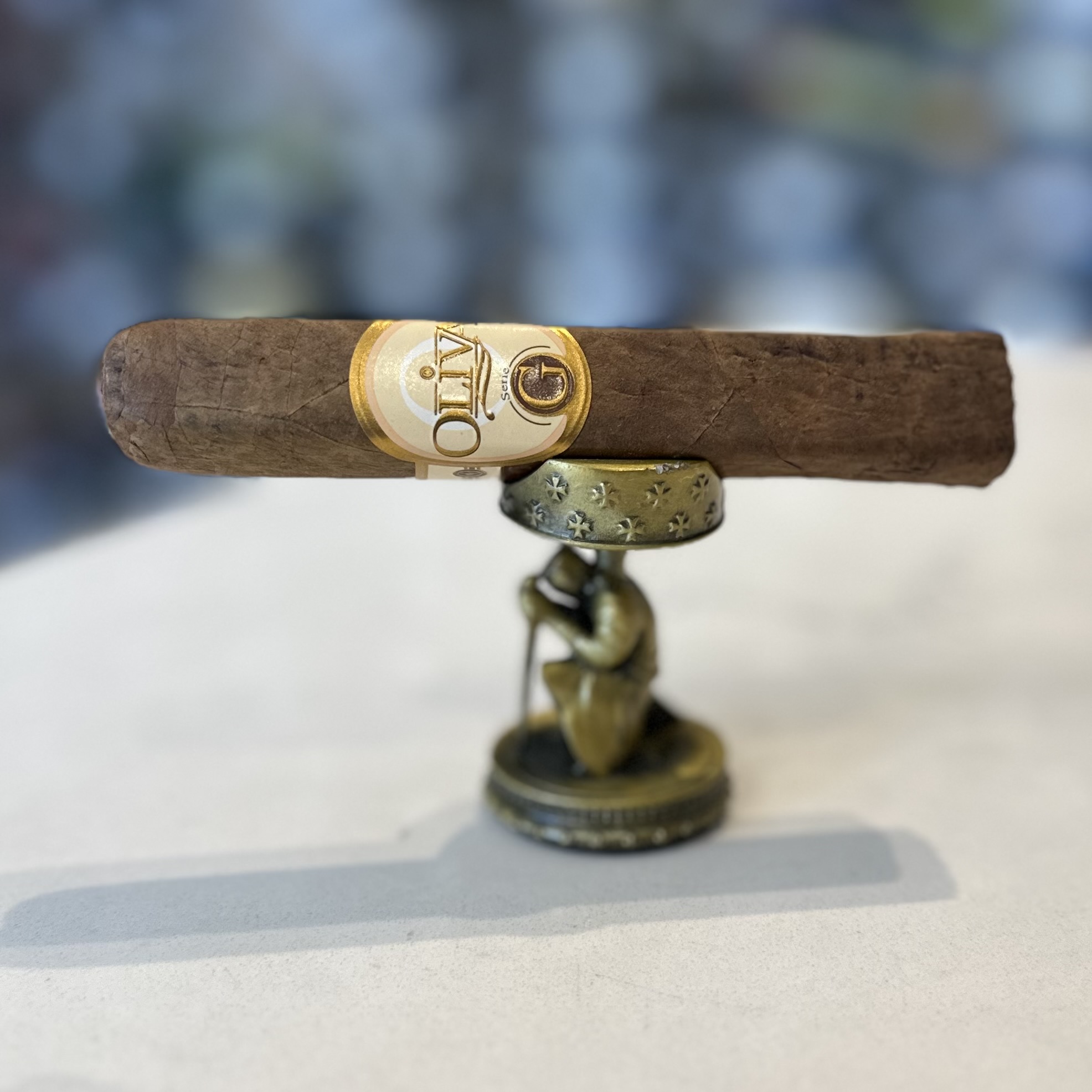 The image displays a premium cigar with a golden and white band labeled "OLIVA" placed horizontally on a small, antique-looking metal stand with a circular base. The stand features decorative elements and a figurine that seems to be supporting the cigar. The background is blurred, focusing the viewer's attention on the cigar and stand, which are centrally positioned in the frame. The cigar appears to have a rich, dark wrapper, suggesting a matured and possibly strong flavor profile, typical of high-quality cigars. The presentation indicates a sense of luxury and tradition, often associated with the ritual of cigar smoking.