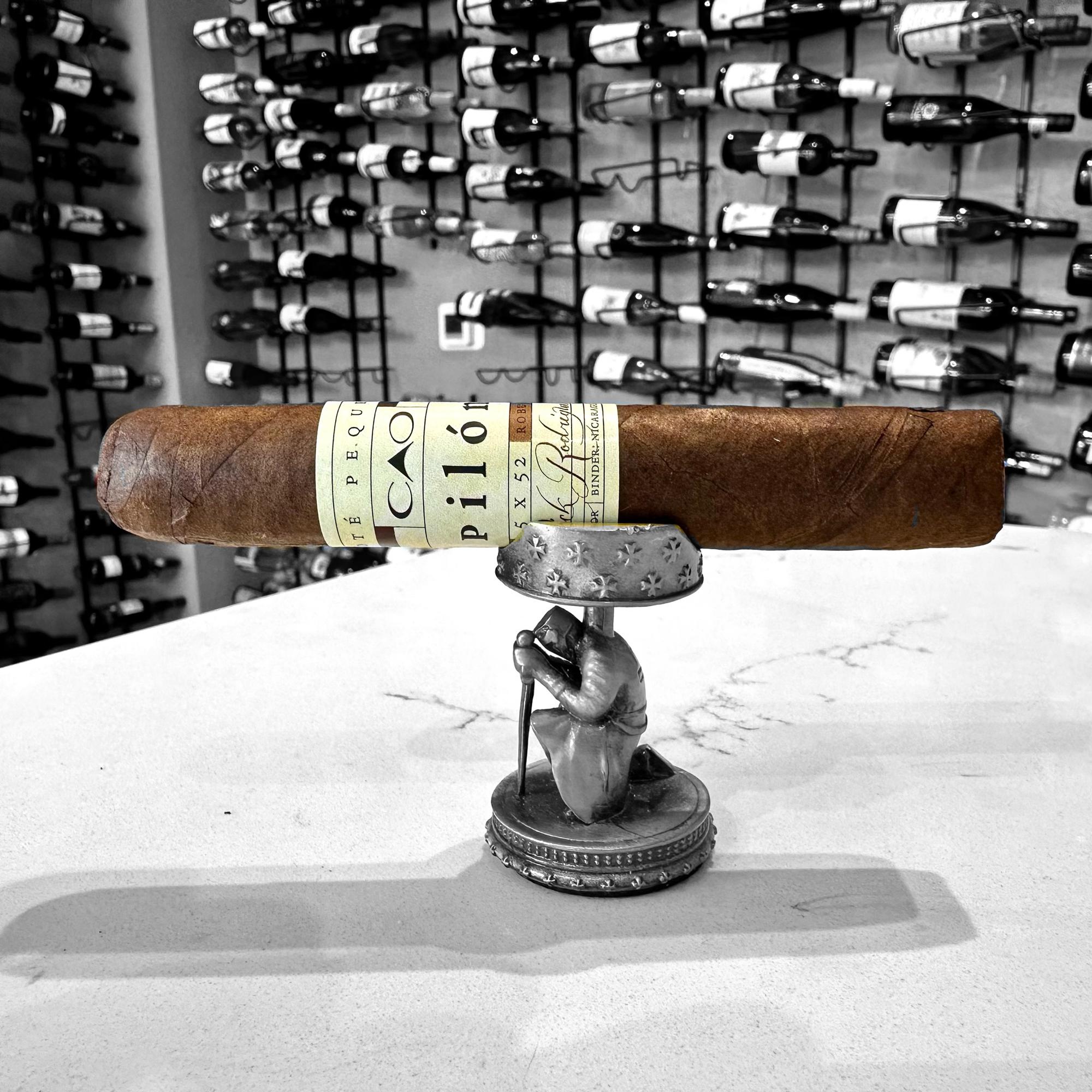 The image shows a premium CAO Pilon cigar resting on a unique cigar stand, which is a miniature sculpture of a figure holding the cigar aloft. The cigar has a rich, brown Ecuador Habano wrapper and is adorned with a distinctive yellow and cream band that features the CAO Pilon branding in elegant typography. The background is a black and white photo of a wall-mounted display of numerous wine bottles, adding an air of sophistication and luxury. The cigar stand and the cigar itself are in full color, creating a striking contrast against the monochromatic backdrop, highlighting the cigar as the central focus of the image.